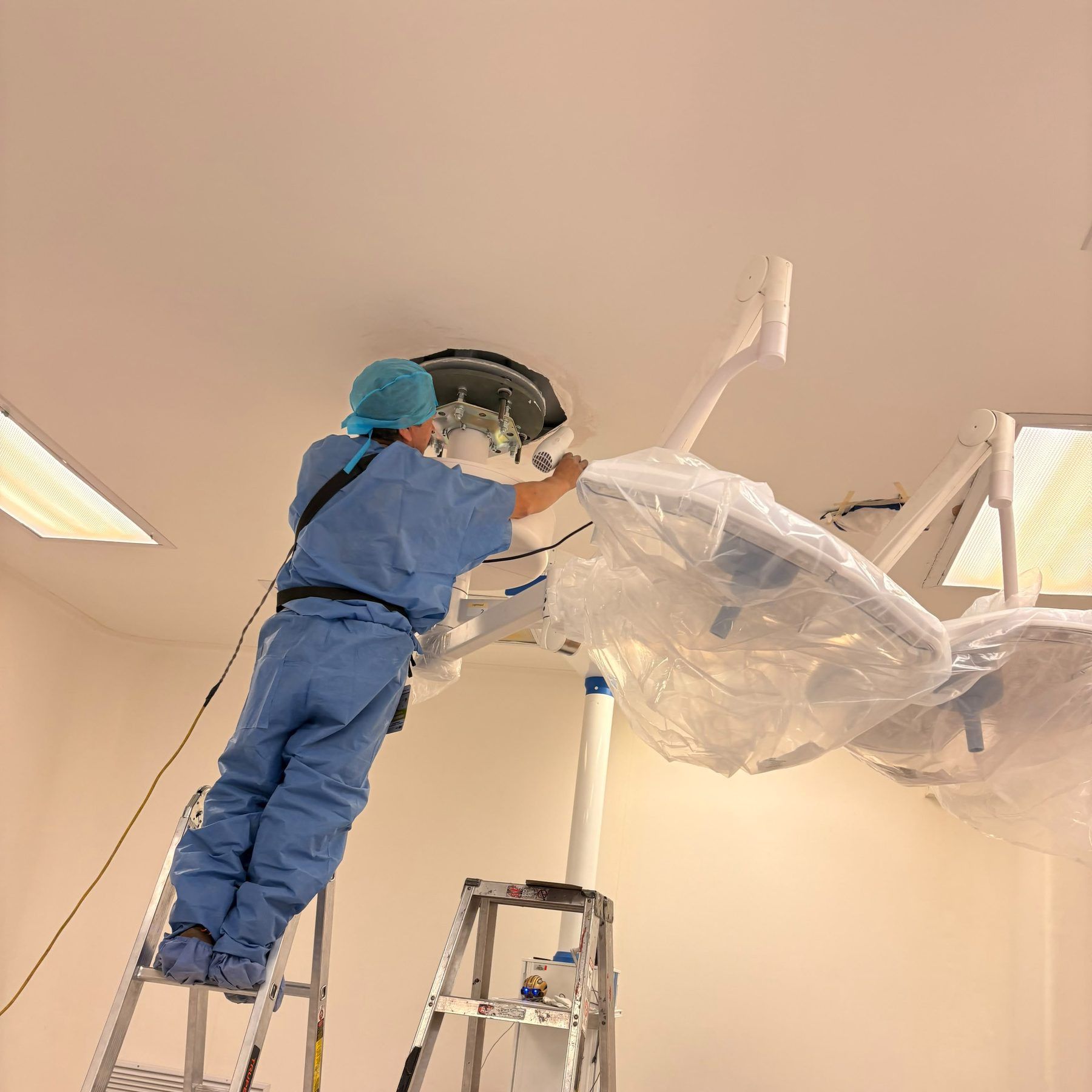 Técnico de BioMed Innovate sobre una escalera instalando una lámpara de cirugía en el techo de un quirófano, con las cúpulas todavía protegidas con funda plástica